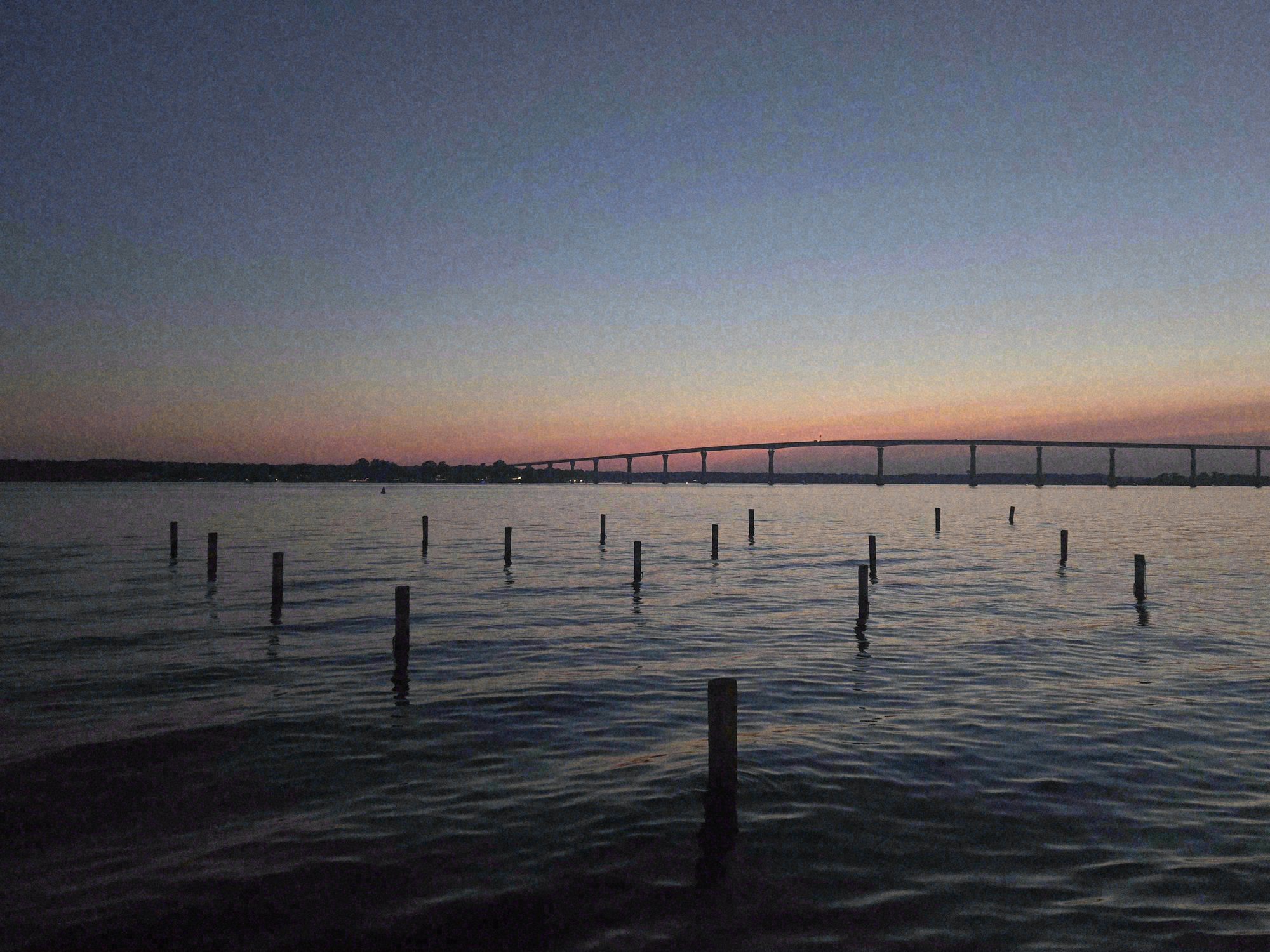 The bridge at sunset in early July. The sky is washed peach above the horizon, deepening to indigo overhead. The pier railing crosses the lower portion of the frame as a quiet horizontal counterweight to the sloping span of the bridge.